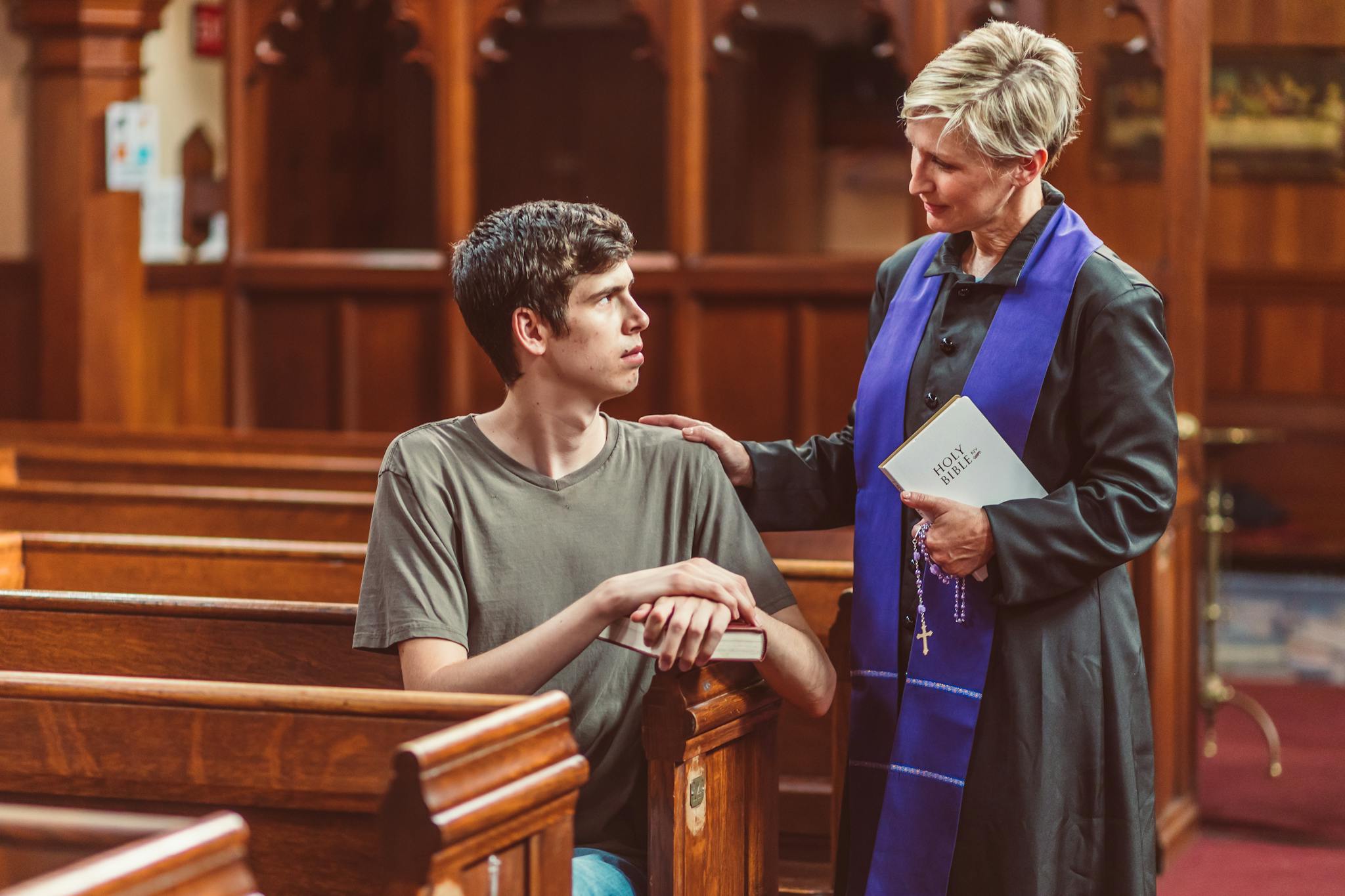 A clergy member comforts a young man in a church pew, discussing faith.