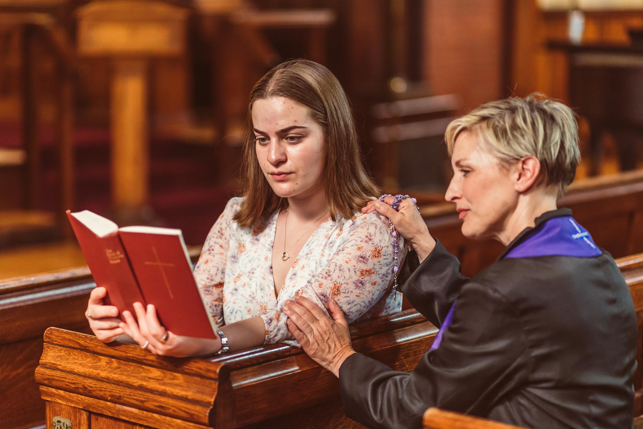 Two women in church, one holding a Bible, engage in thoughtful spiritual reading and prayer.